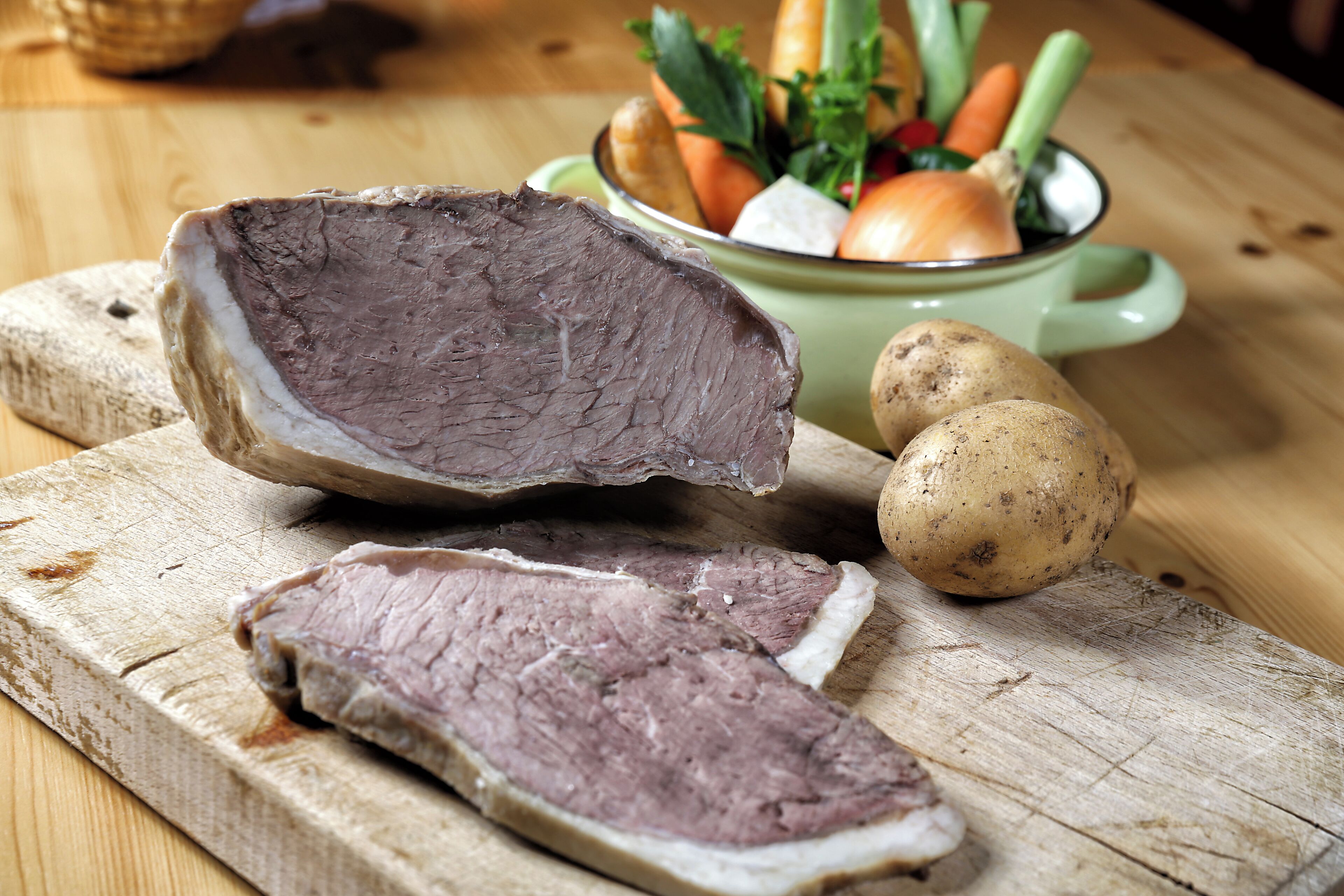 Ein großes Stück Fleisch, geschnittenes Rindfleisch und zwei Kartoffeln auf einem Holztisch.