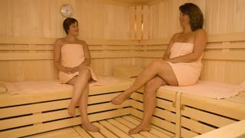 Dos personas sentadas en una sauna con bancos de madera y un reloj visible en la pared.