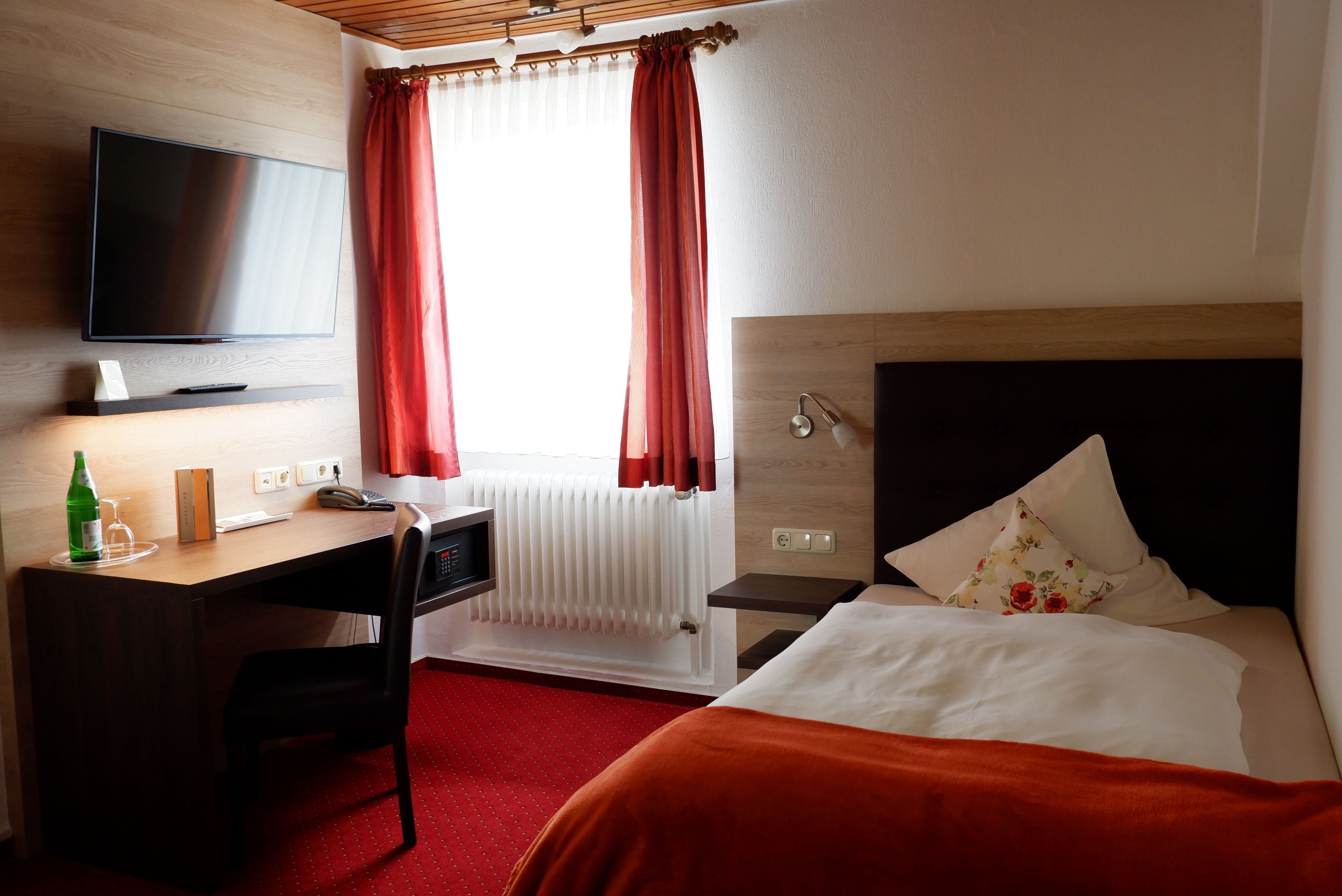 Habitación de hotel con cama, escritorio, televisión, ventana con cortinas, radiador y mesita de noche.