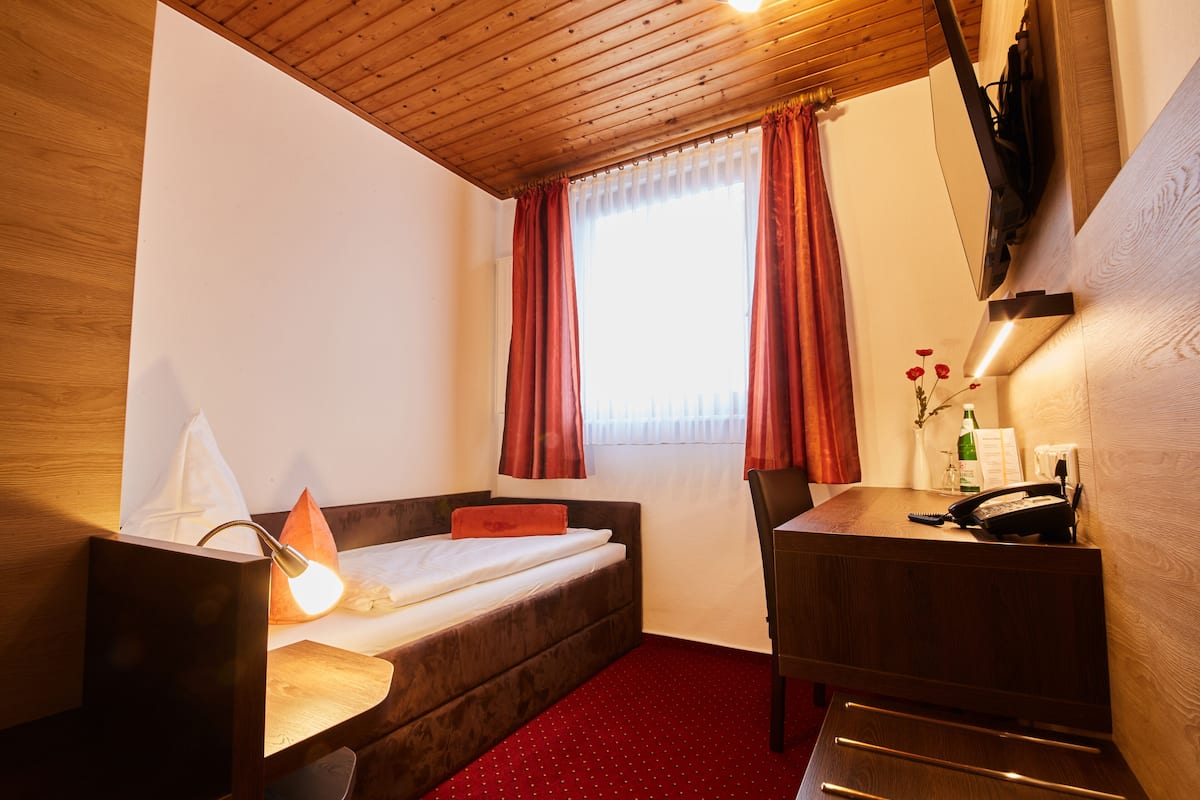 A compact hotel room with a single bed, a desk, and a chair. A lamp is on the desk, and a bottle of water is on the nightstand.