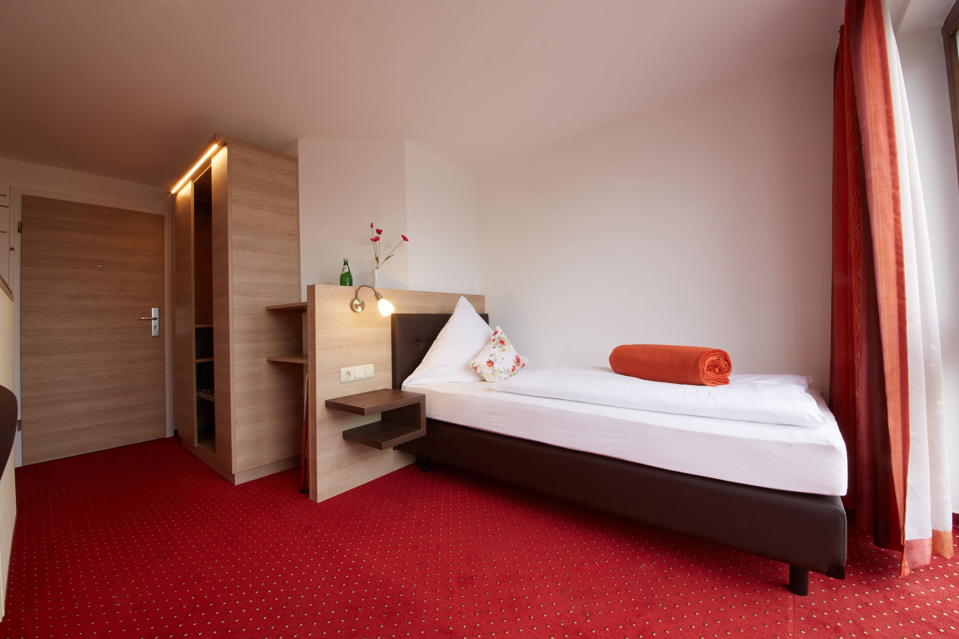 Una camera d'albergo con un letto, un armadio in legno, un piccolo tavolo con una lampada e un tappeto rosso.