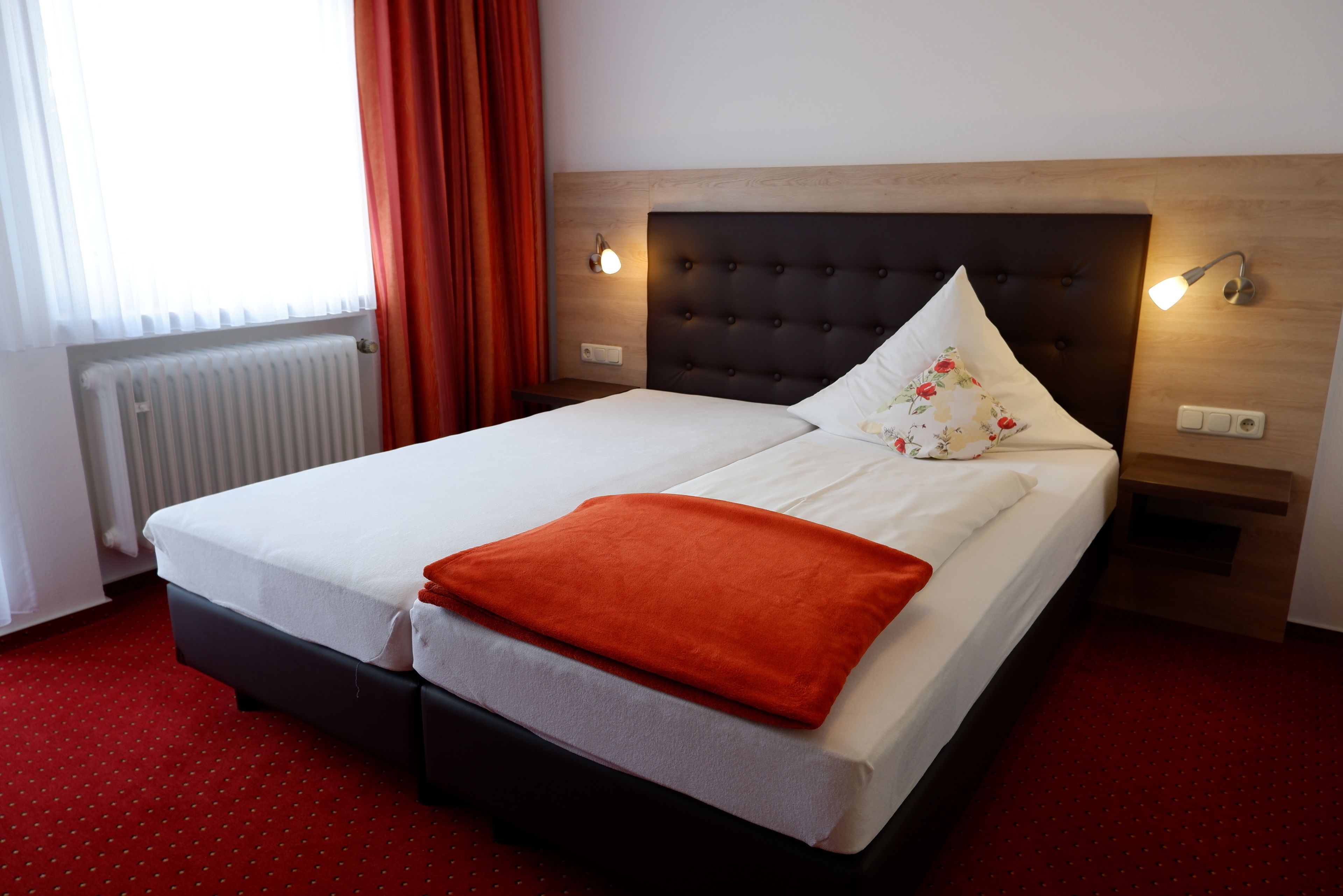 A neatly made bed with a white and orange blanket, a wooden headboard, and two wall-mounted reading lamps.