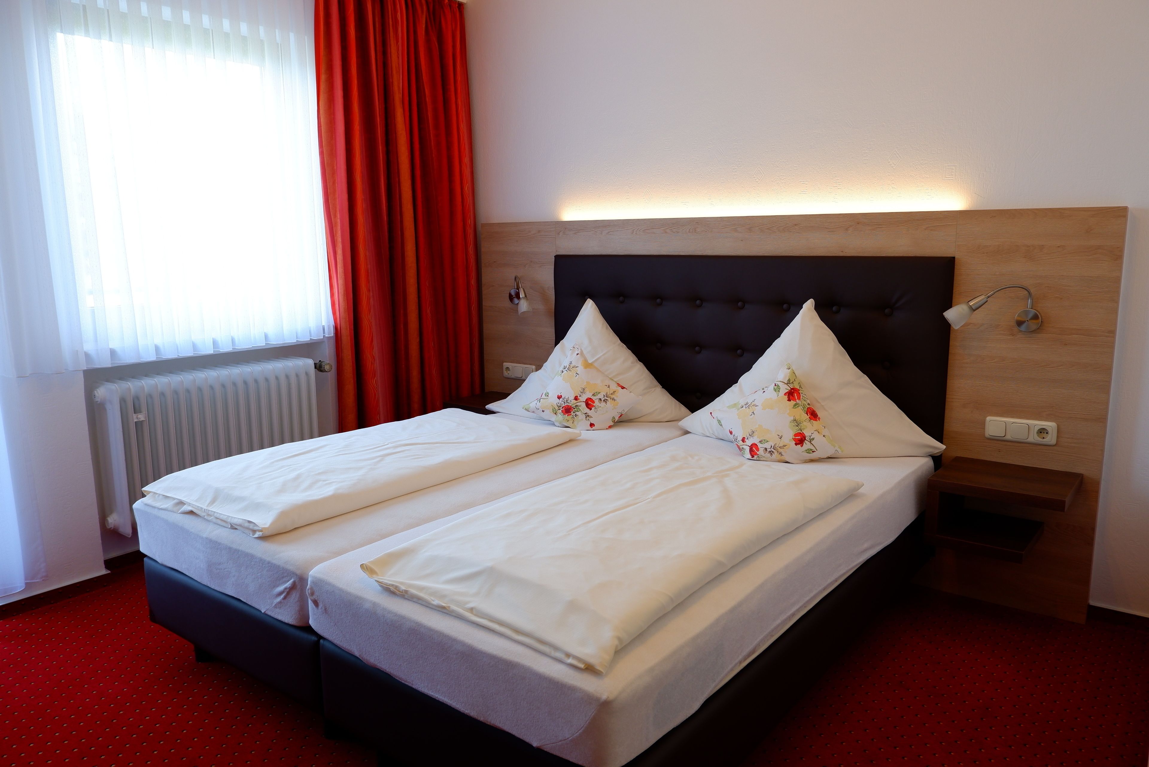 A double bed with white linens and floral pillows, a wooden headboard, a bedside table with a lamp, and a red carpeted floor.