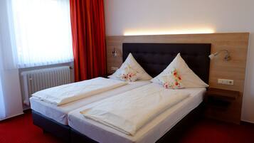 A double bed with white linens and floral pillows, a wooden headboard, a bedside table with a lamp, and a red carpeted floor.