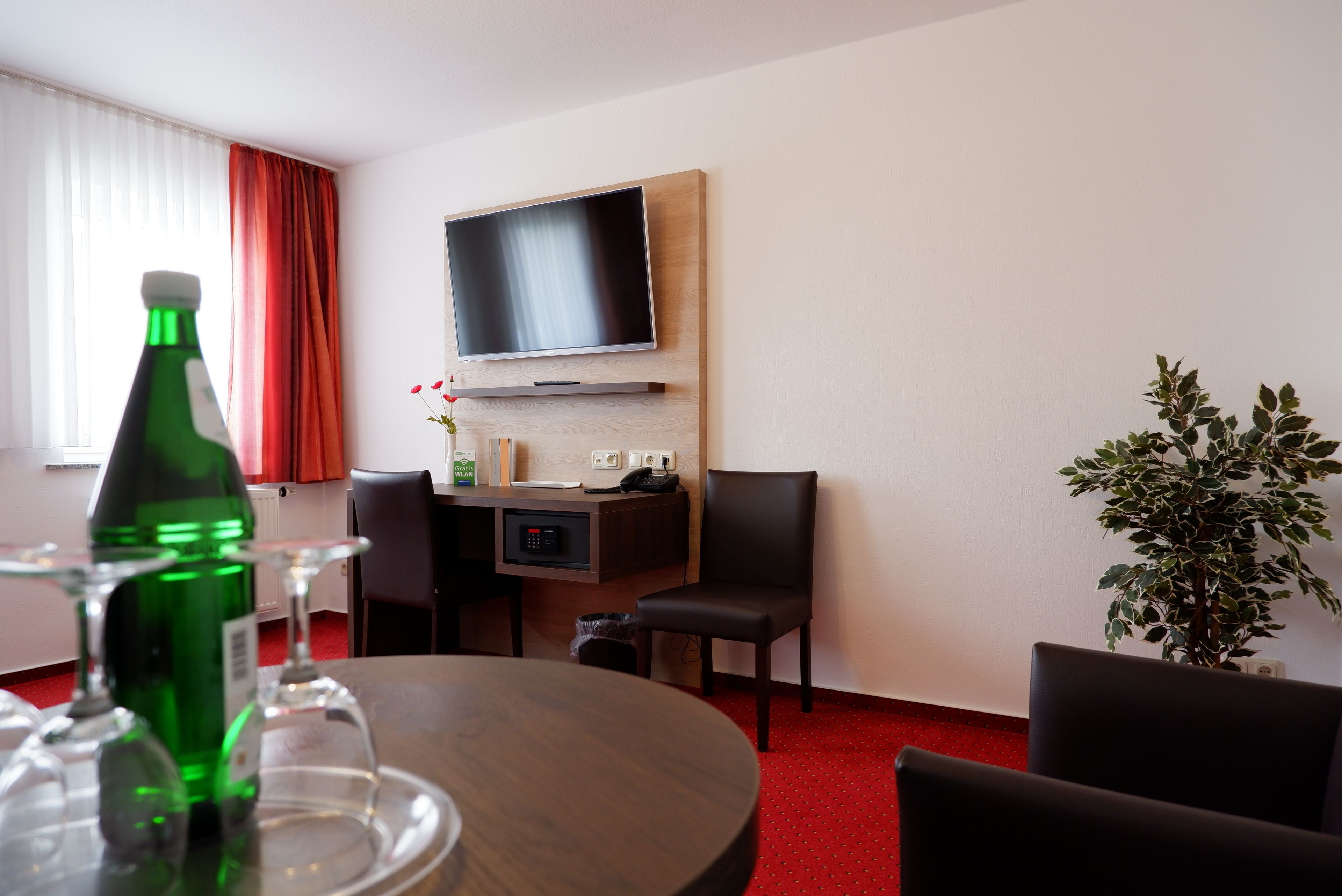 Ein Hotelzimmer mit einem runden Esstisch, einem flachbildfernseher an der Wand, einer Topfpflanze und einem roten Teppichboden.