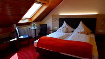 A room with a sloped ceiling, a bed with white bedding and red accents, a small table with a chair, and a radiator.
