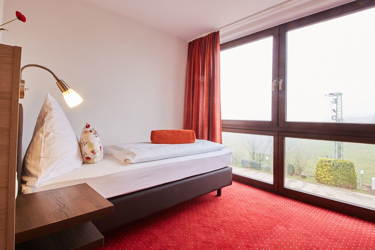 A hotel room with a bed, a nightstand, a lamp, a window with curtains, and a view of the outside.