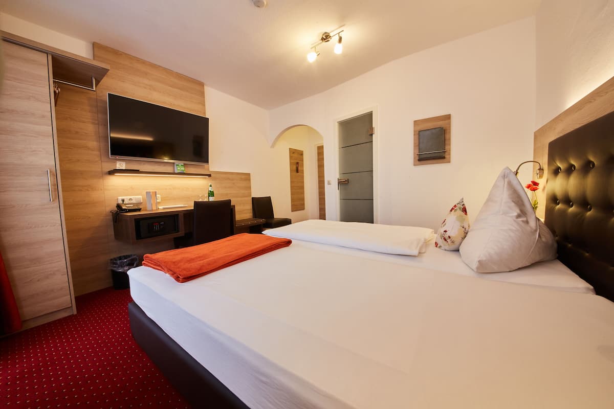 A hotel room with a bed, a desk with a computer, a TV mounted on the wall, and a red carpet.