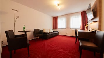 Habitación de hotel con alfombra roja, un sofá, dos sillones, una mesita, un televisor montado en la pared y una ventana con cortinas.