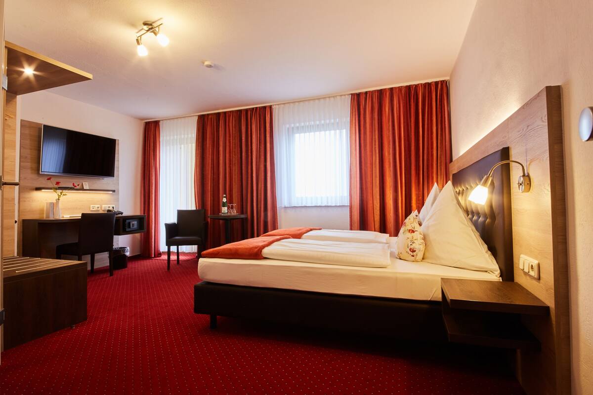 A hotel room with a bed, desk, chair, TV, and a red carpeted floor.