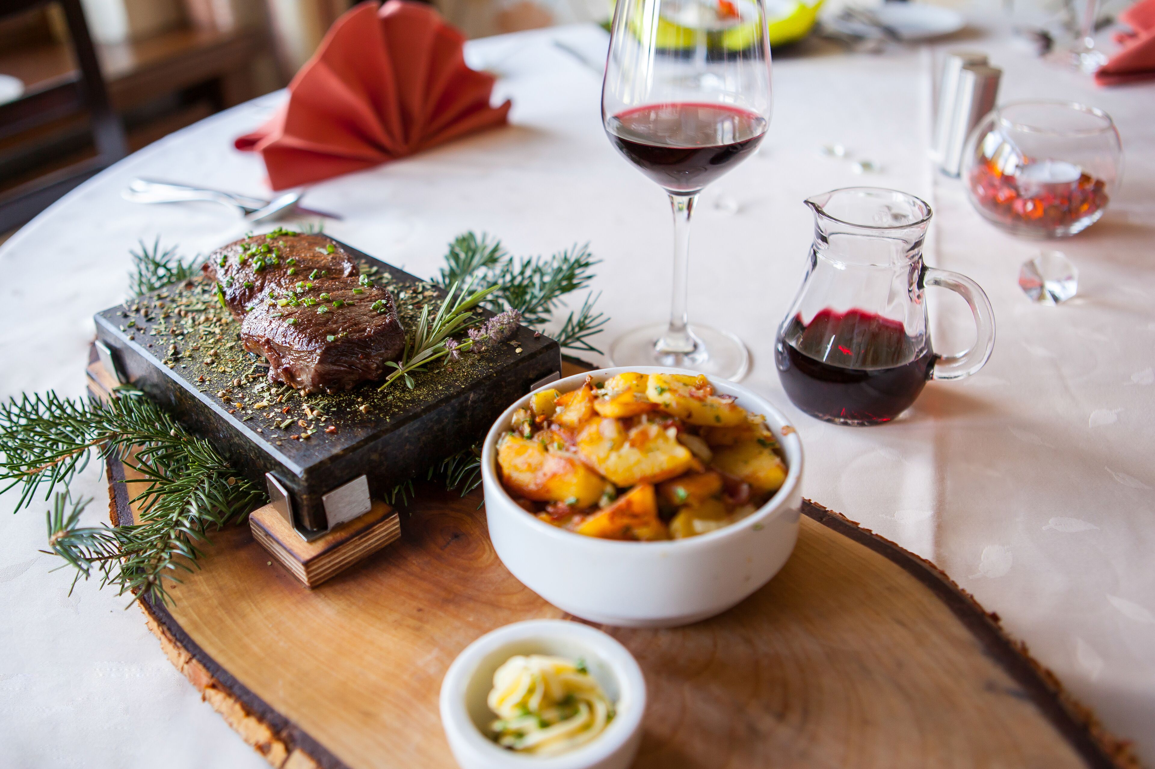 Una mesa de comedor con un plato de carne asada como centro, un tazón de papas y una copa de vino tinto.