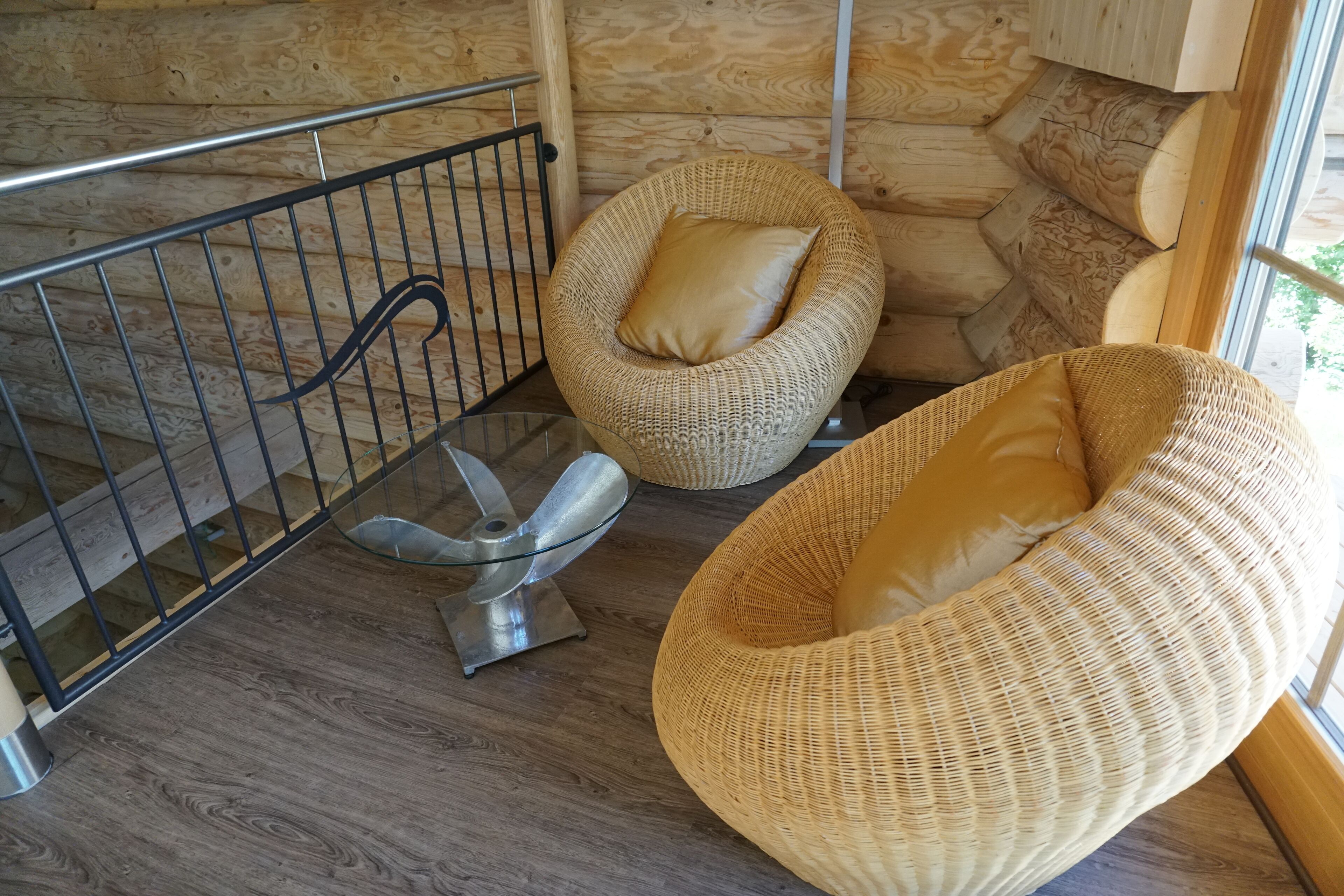 A wooden interior with a metal railing, two wicker chairs with cushions, and a glass table.