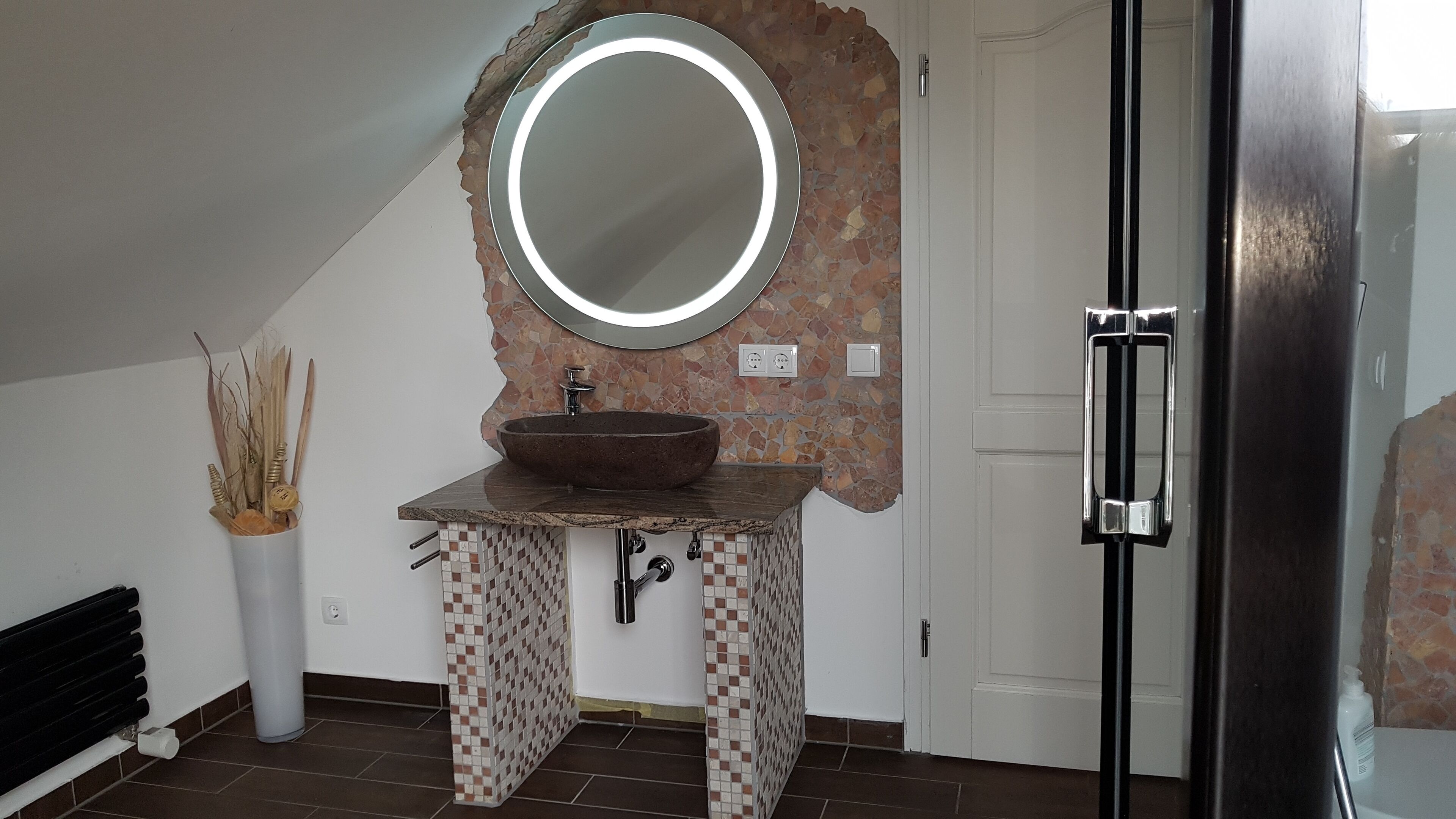 Een badkamer met een stenen wasbak, een ronde spiegel en een moderne deur.
