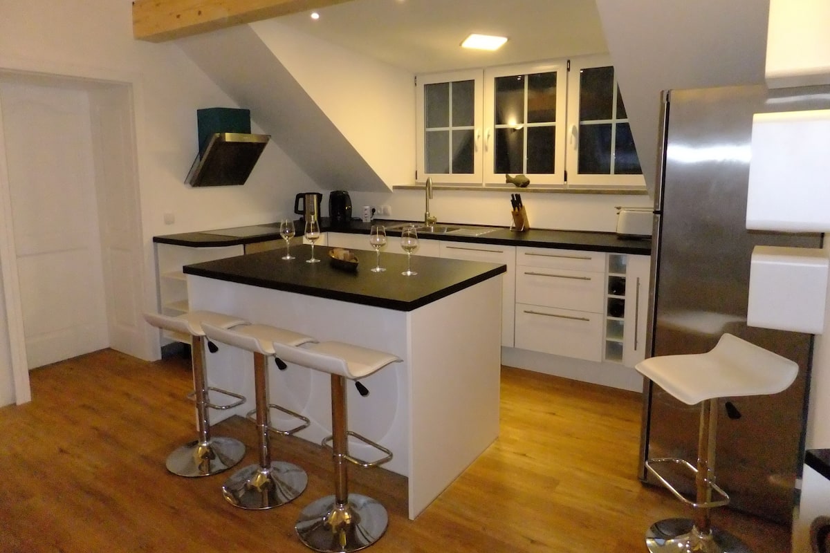 A modern kitchen with a central island, bar stools, a stainless steel refrigerator, and a wooden ceiling.