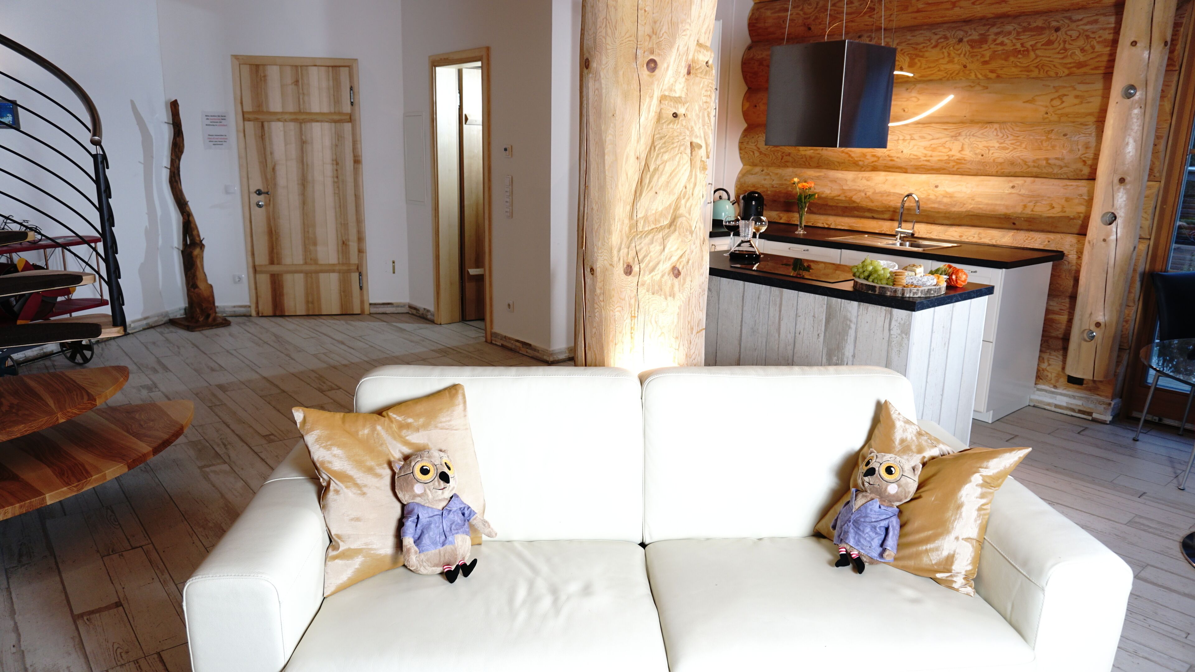 Een gezellige woonkamer met een witte bank, twee teddyberen, een houten salontafel en een keukenhoek met een hanglamp.
