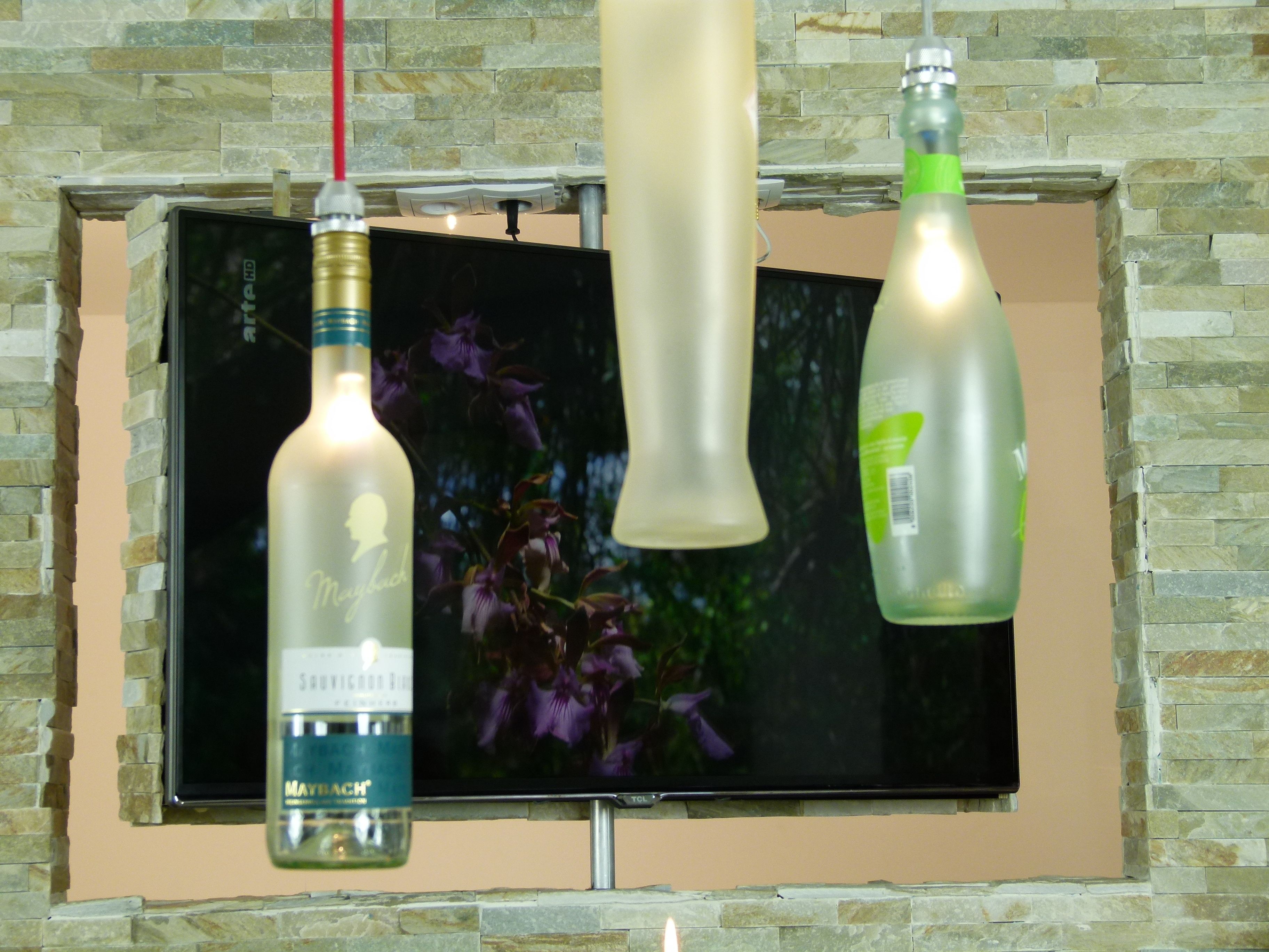 A television mounted on a stone wall with three bottles hanging above it.
