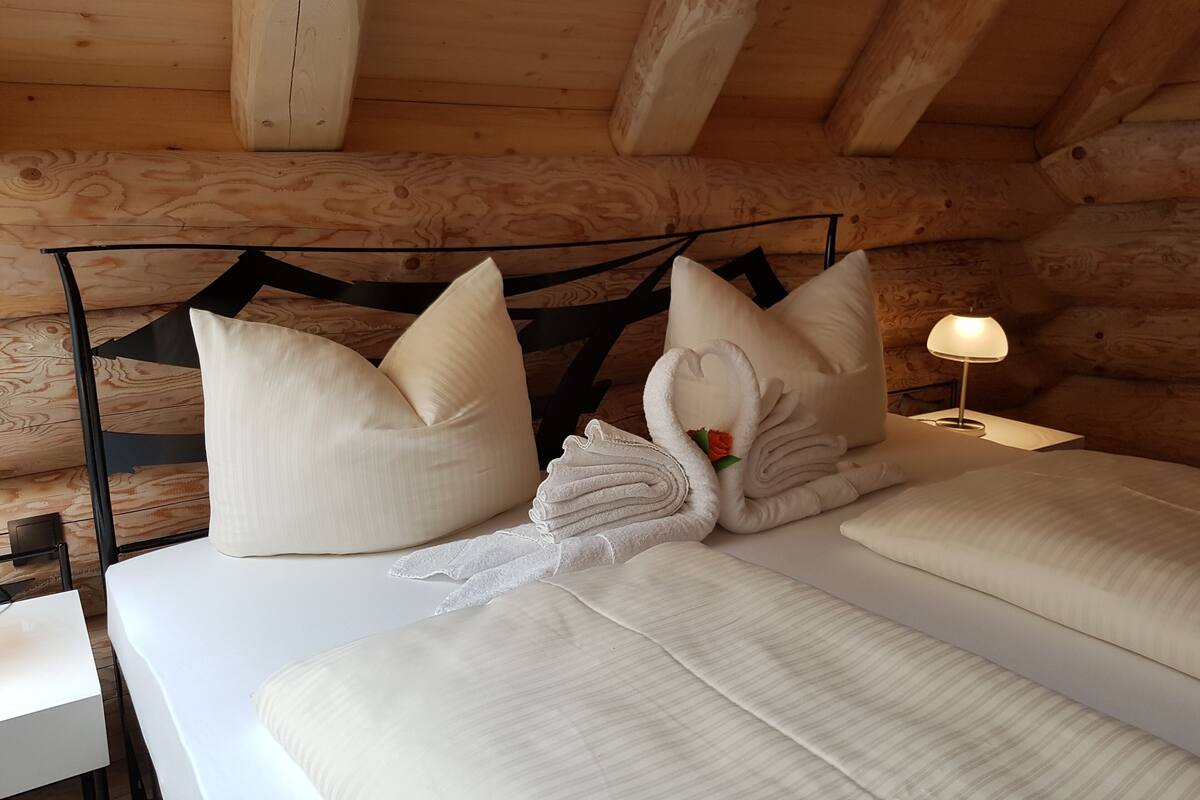 A cozy wooden cabin room with a bed, pillows, a swan-shaped towel, and a nightstand with a lamp.