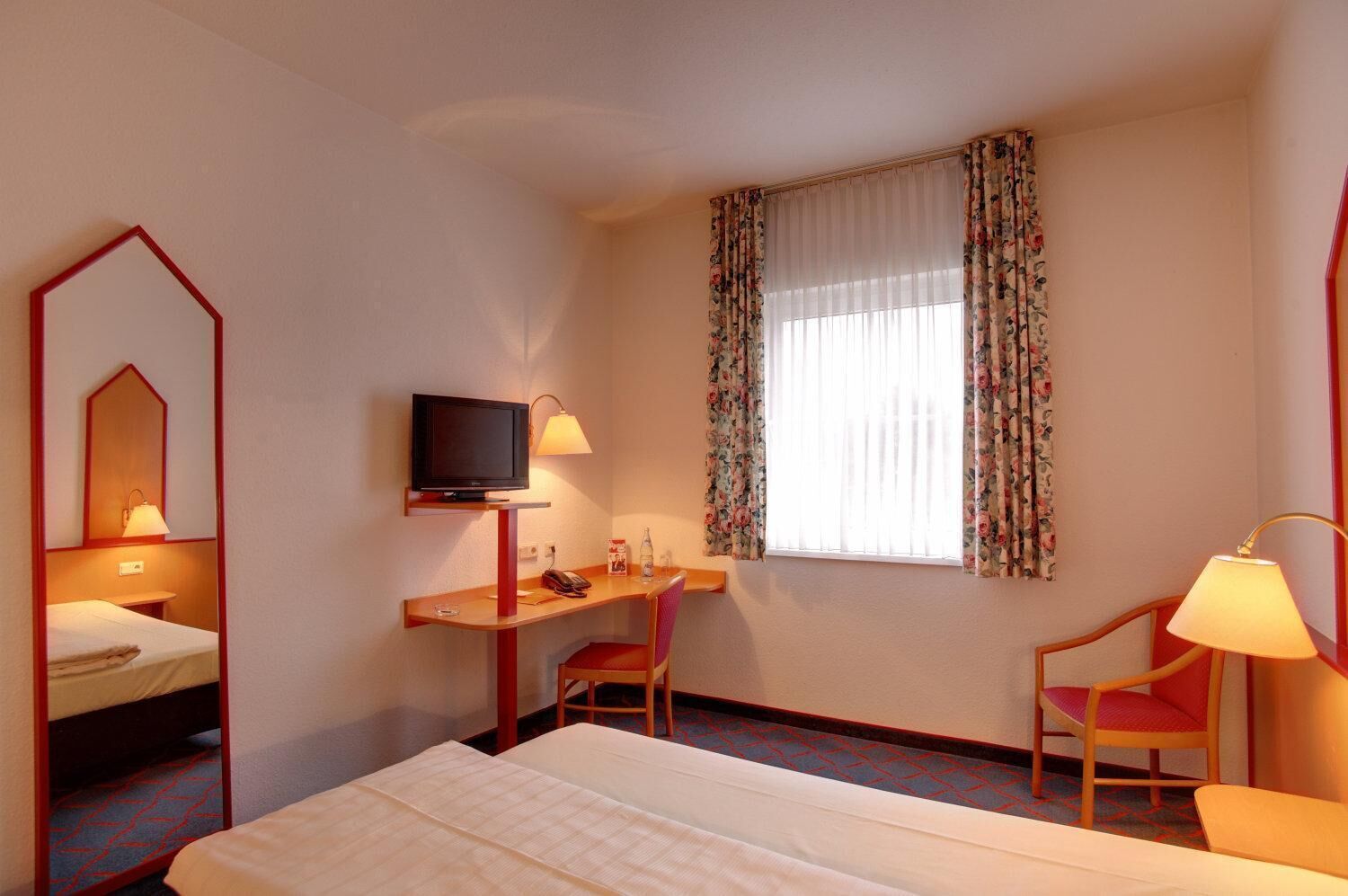 A hotel room with a bed, a desk with a television, a chair, and a window with floral curtains.