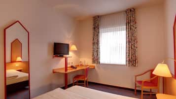 A hotel room with a bed, a desk with a television, a chair, and a window with floral curtains.