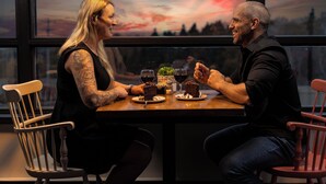 Two people seated at a table with a sunset view through a window, enjoying a meal with wine and desserts.