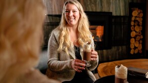 A person holding a glass of a dark beverage, possibly coffee, in a cozy indoor setting with a fireplace and a stack of logs in the background.
