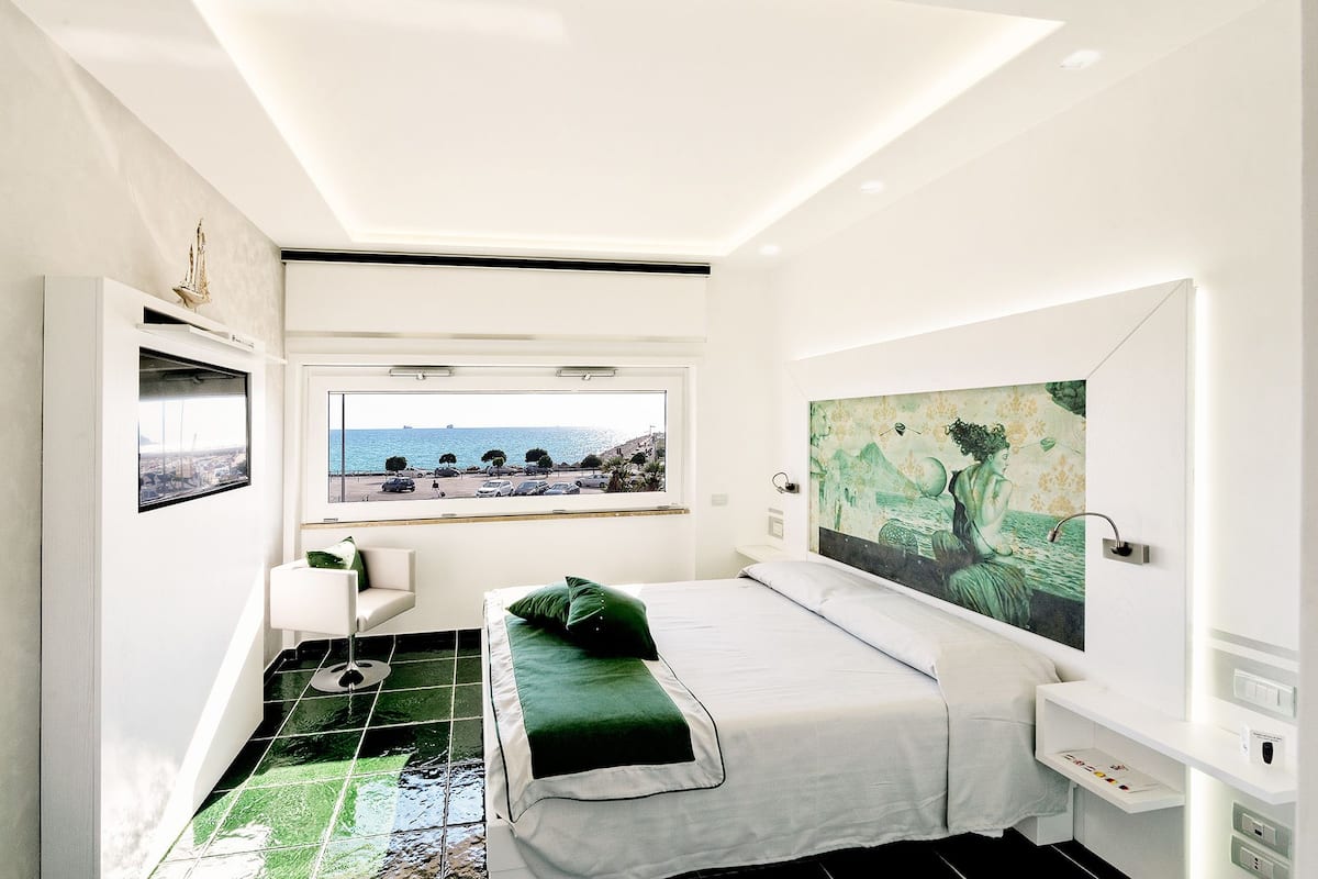 A modern bedroom with a bed, a bench, a window with a view of the sea, and a painting on the wall.