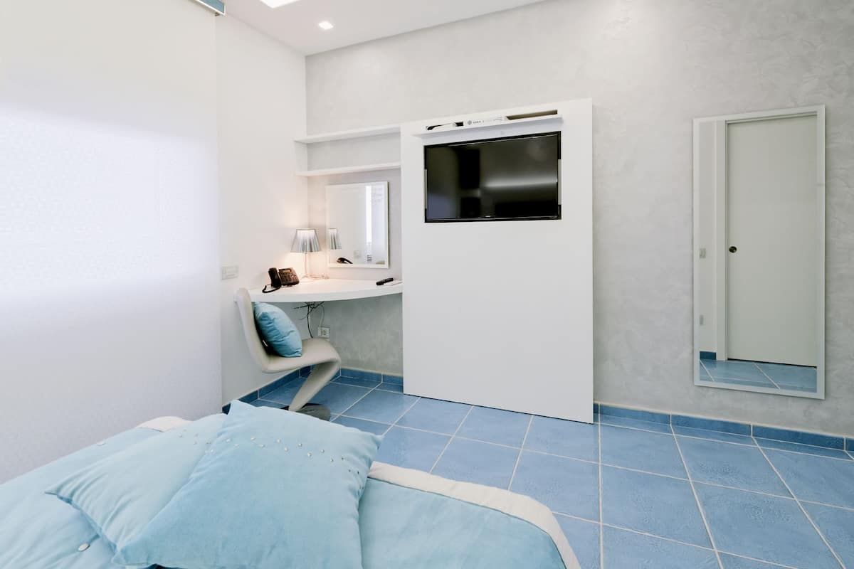 A compact hotel room with a bed, a desk with a computer, a chair, a small table with a lamp, and a door.
