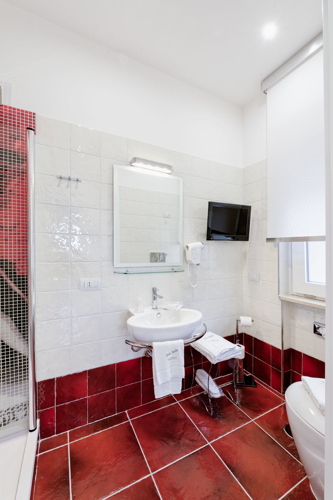 Une salle de bain avec un lavabo blanc, un miroir, un téléviseur fixé au-dessus du lavabo et un espace douche dont les murs sont recouverts de carreaux rouges.