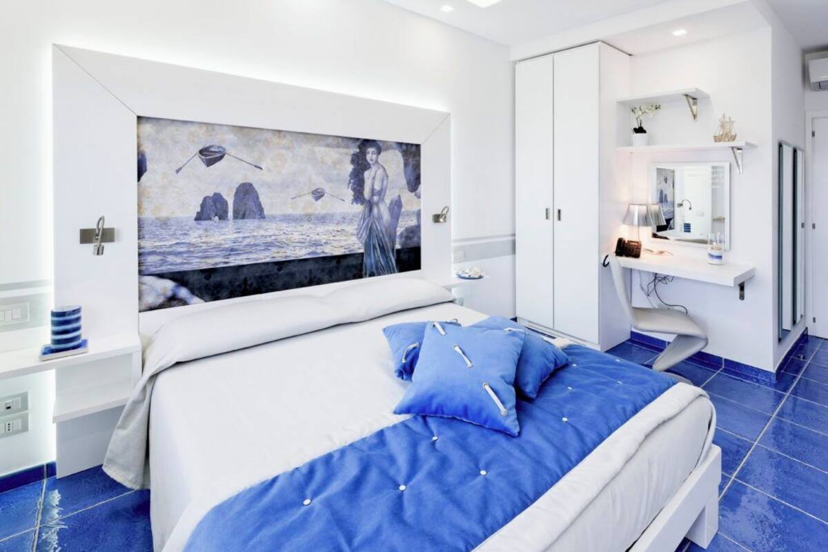 A modern bedroom with a large bed, a painting of a seascape, a white wardrobe, and a vanity with a mirror.