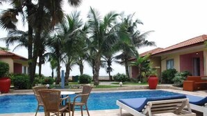 A pool area with lounge chairs and a table, surrounded by palm trees and a building with a red roof. - Résidence Saint-Jacques Bord de Mer (Pointe-Noire)