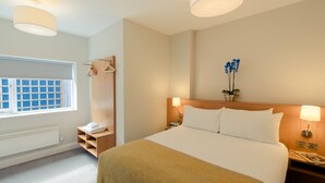A hotel room with a large bed, bedside lamps, a window with blinds, a wooden wardrobe, and a small potted plant. - PREMIER SUITES Reading (Reading)