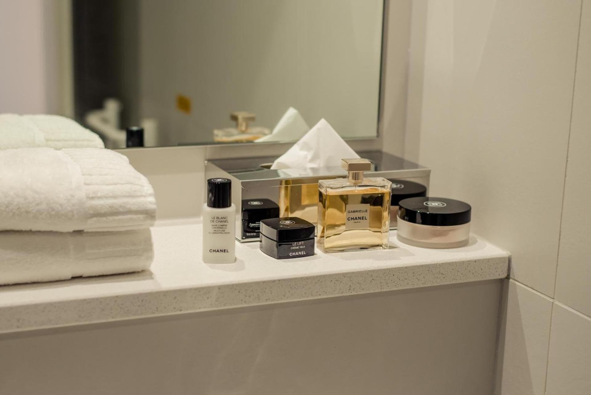 En badeværelsesvask med toiletartikler, heriblandt en flaske Chanel Le Blanc de Chanel, en parfumeflaske og et pudder.