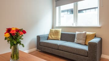 Ein graues Sofa mit zwei Kissen, eine transparente Vase mit Blumen und ein Fenster mit Jalousien.