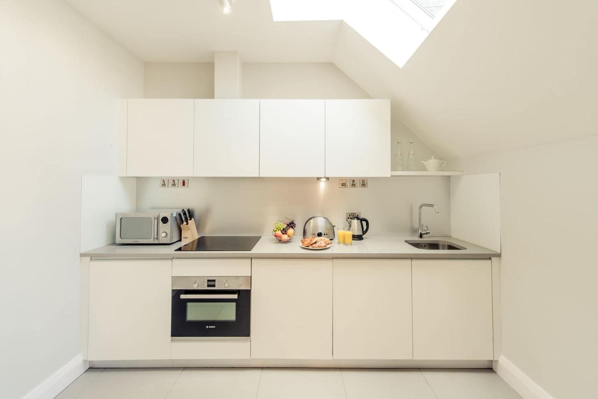 A modern kitchen with white cabinetry, a built-in oven, a microwave, a sink, and a kettle.