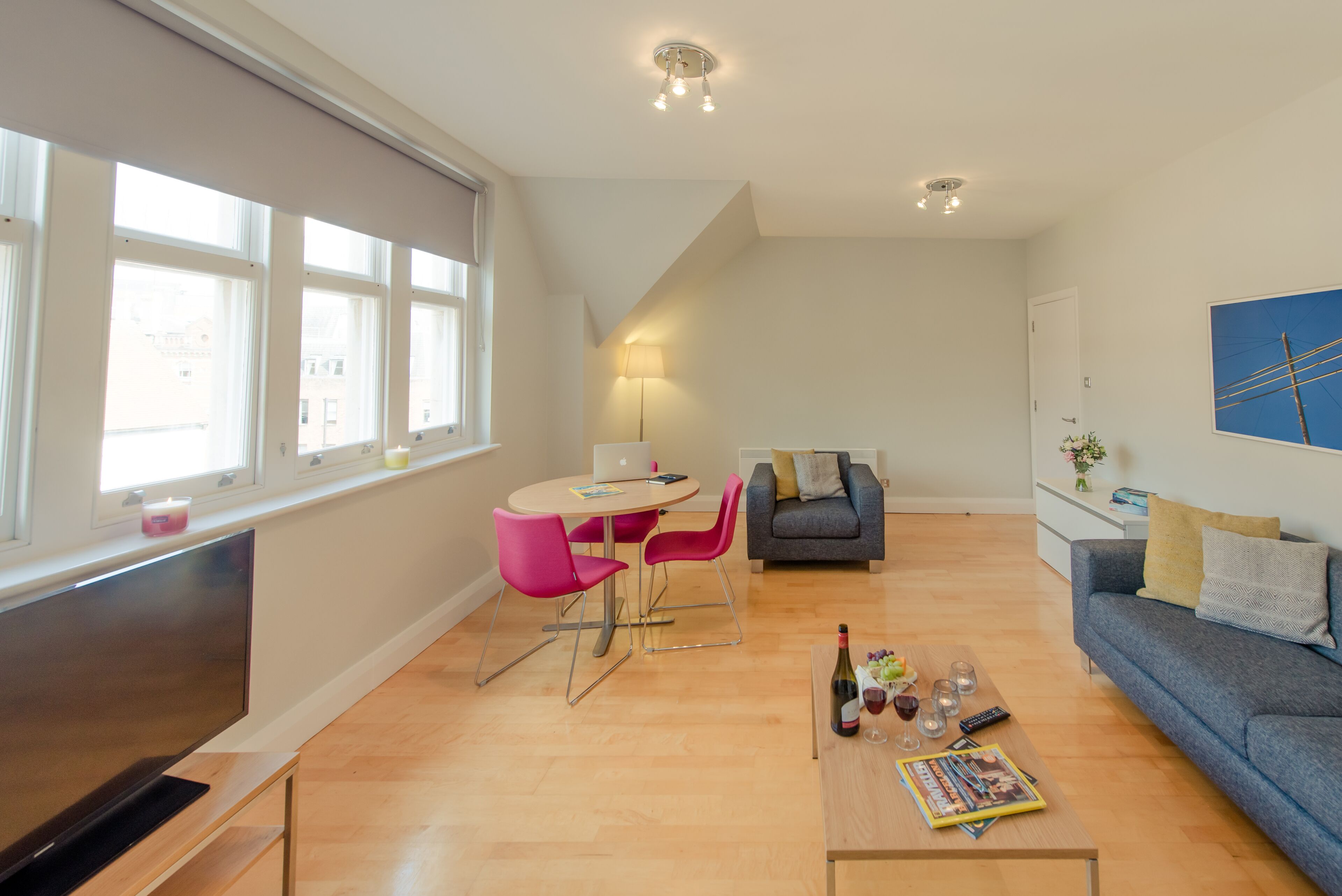 A modern living room with a flat-screen TV, a dining area with pink chairs, a sofa, and a coffee table with a magazine and wine glasses.