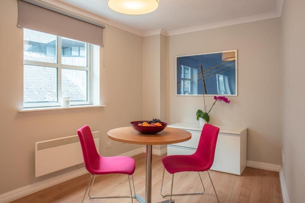 A room with a round table, two pink chairs, a bowl of fruit, a window, and a painting on the wall.