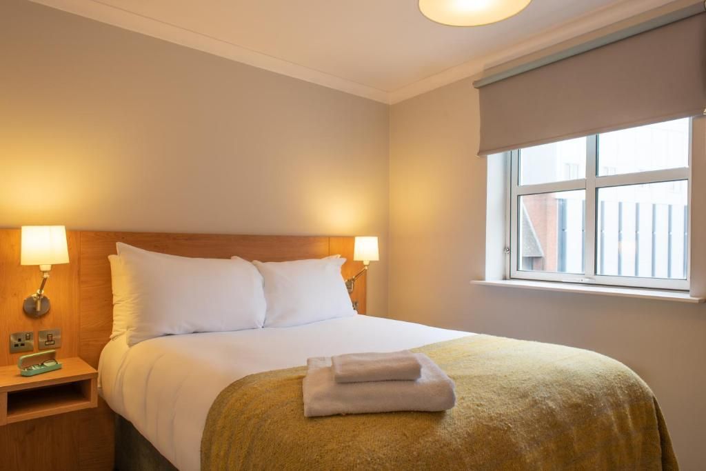 A hotel room with a bed, bedside lamps, a window with a view, and a neatly made bed with a folded towel.