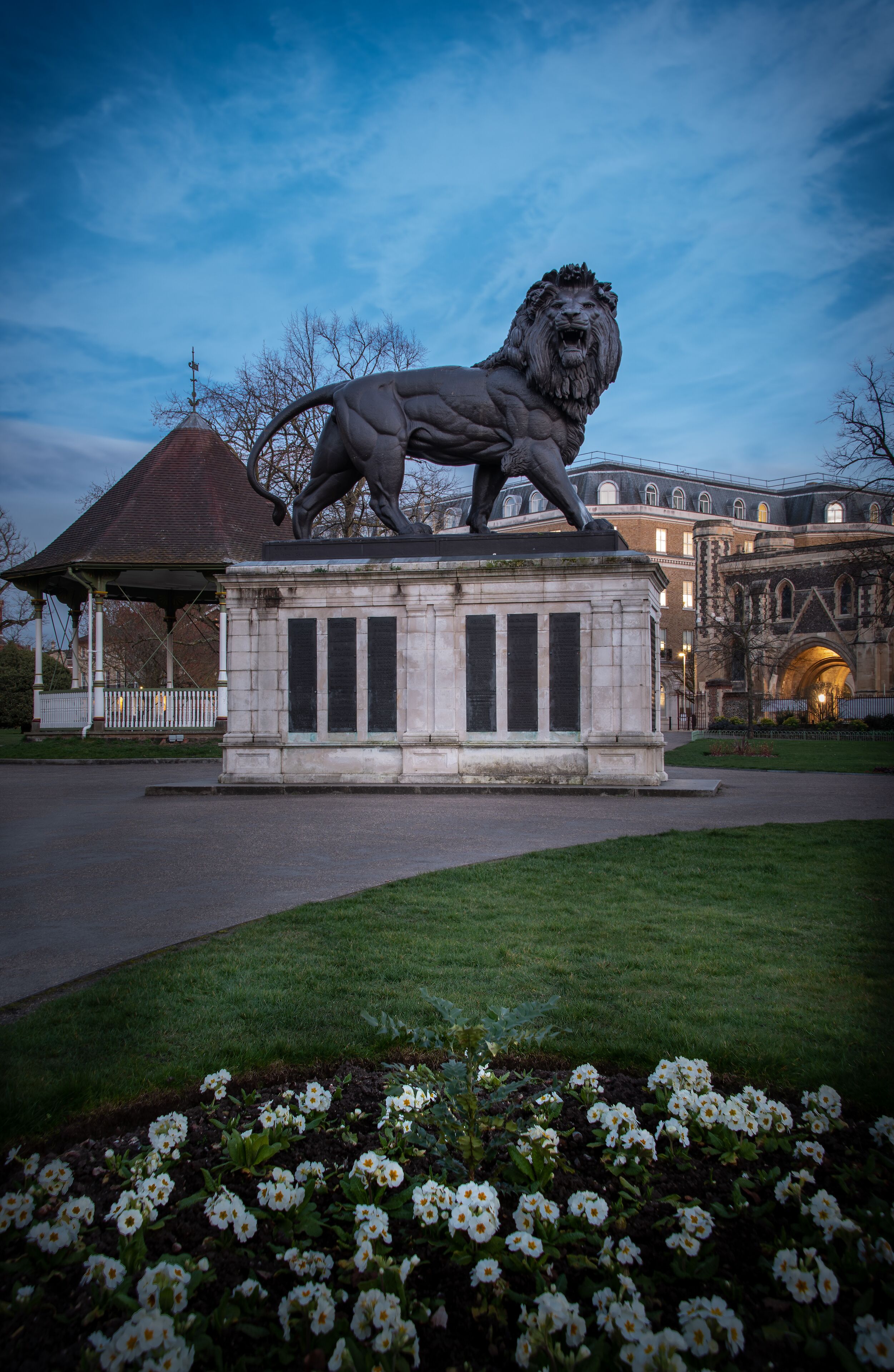 A lion statue on a pedestal in a park with a gazebo and a building in the background.
