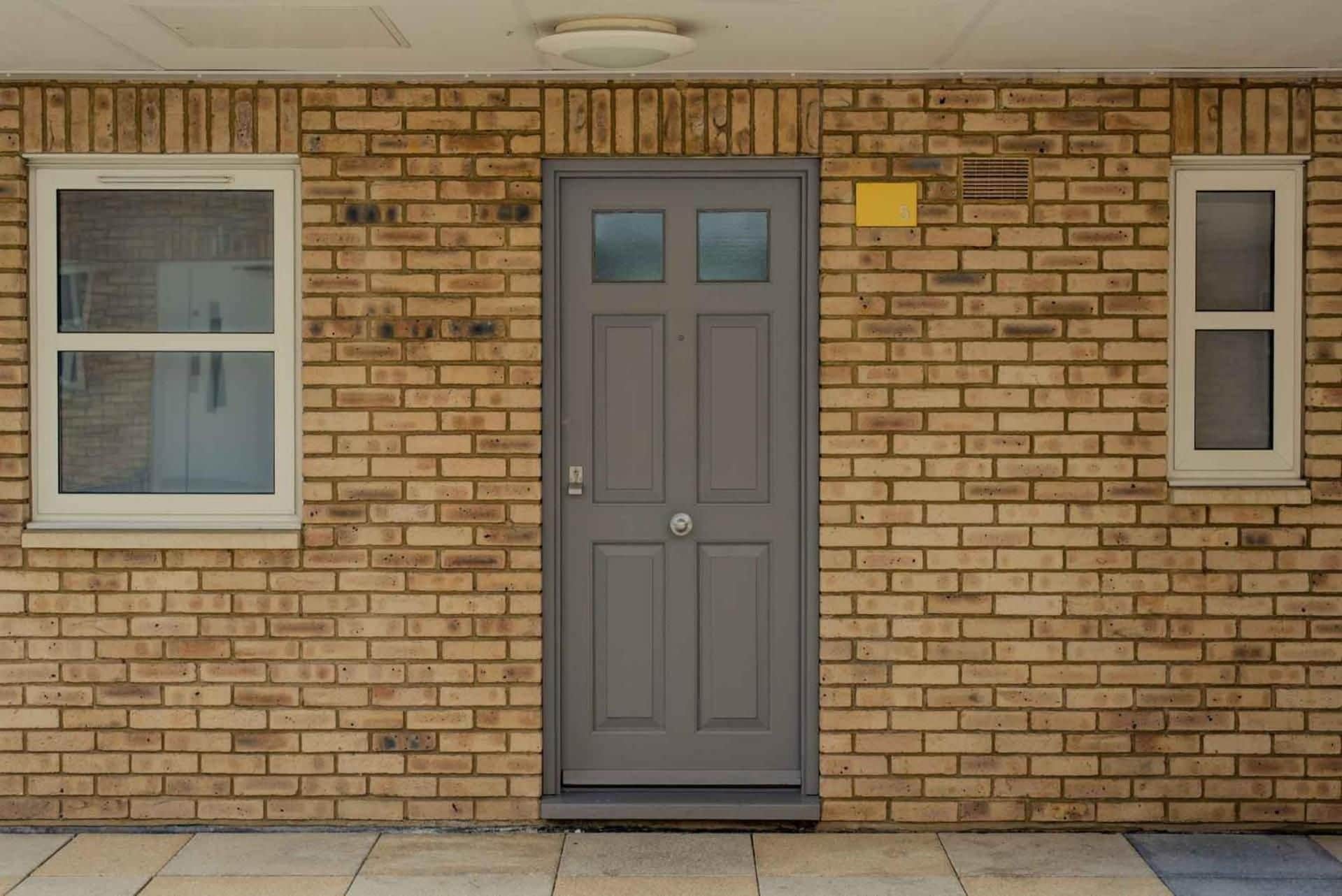 Un bâtiment en briques avec une porte centrale, deux fenêtres de chaque côté et une petite grille d’aération sur le mur de droite.