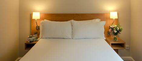 Una cama bien hecha con sábanas blancas, dos lámparas de noche y una mesita de noche con un florero.
