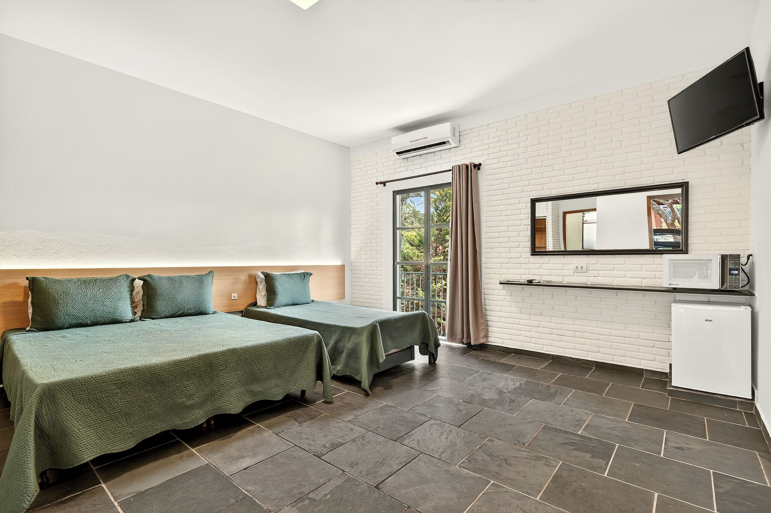 A hotel room with two beds, a kitchenette, a flat-screen TV, and a large window with curtains.