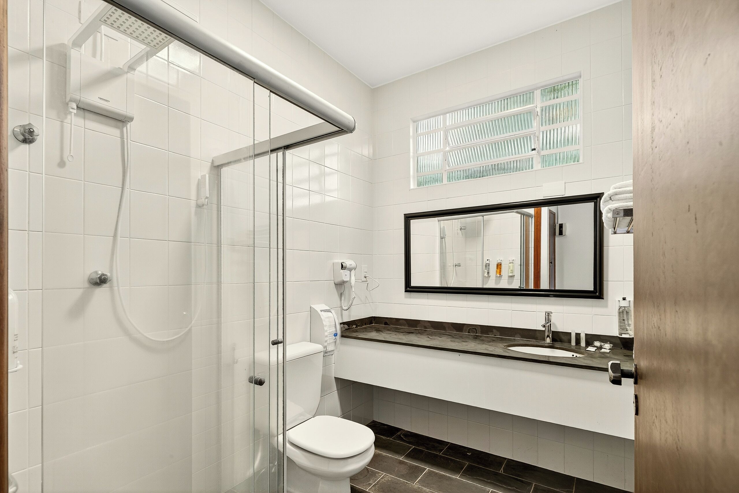A modern bathroom with a glass shower enclosure, a toilet, a sink with a mirror, and a window with blinds.