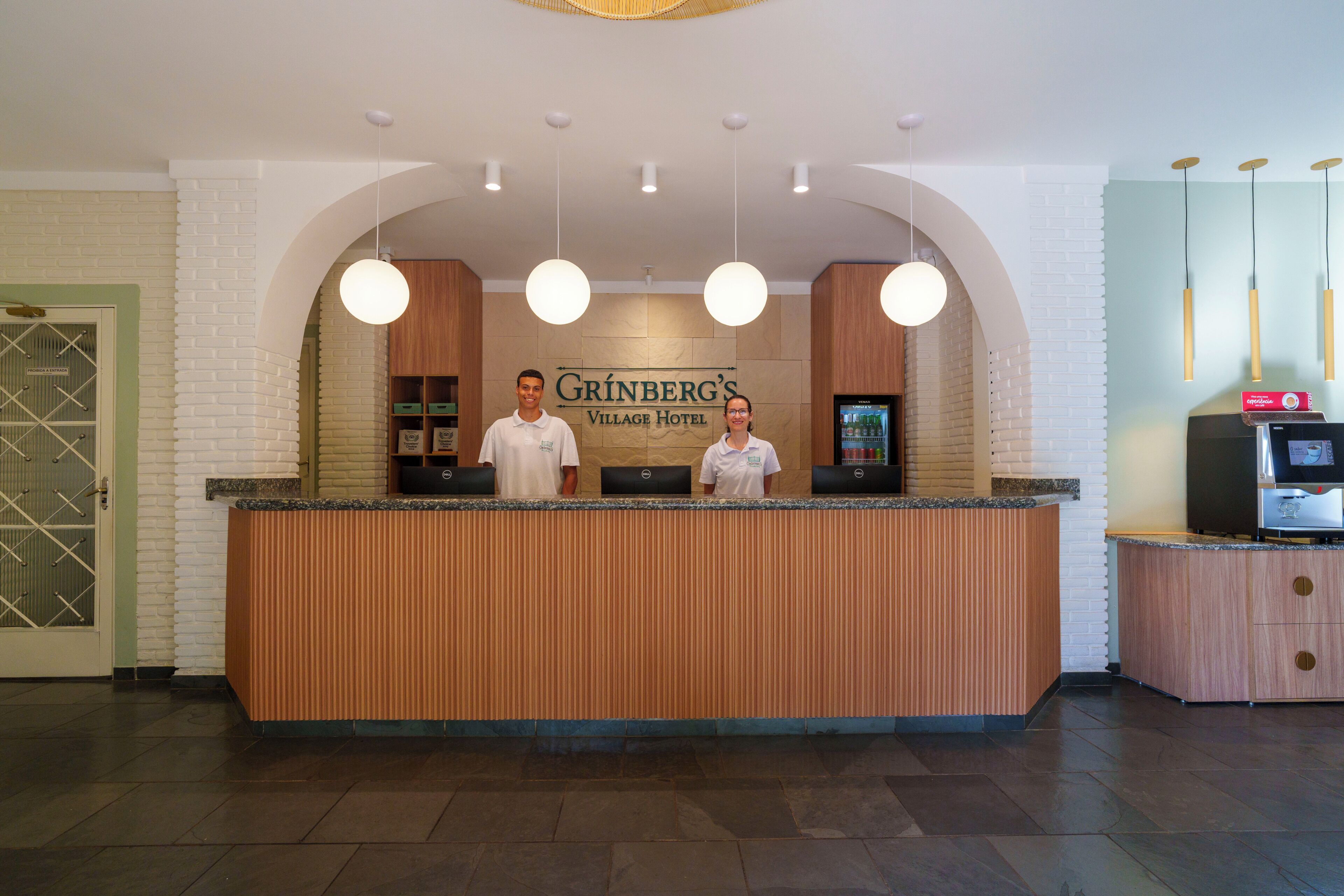 A reception desk at Grinberg's Village Hotel with a wooden finish, a sign above, and a refrigerator to the right.