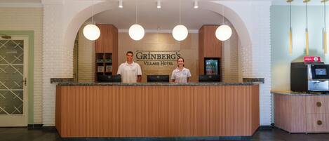 A reception desk at Grinberg's Village Hotel with a wooden finish, a sign above, and a refrigerator to the right.