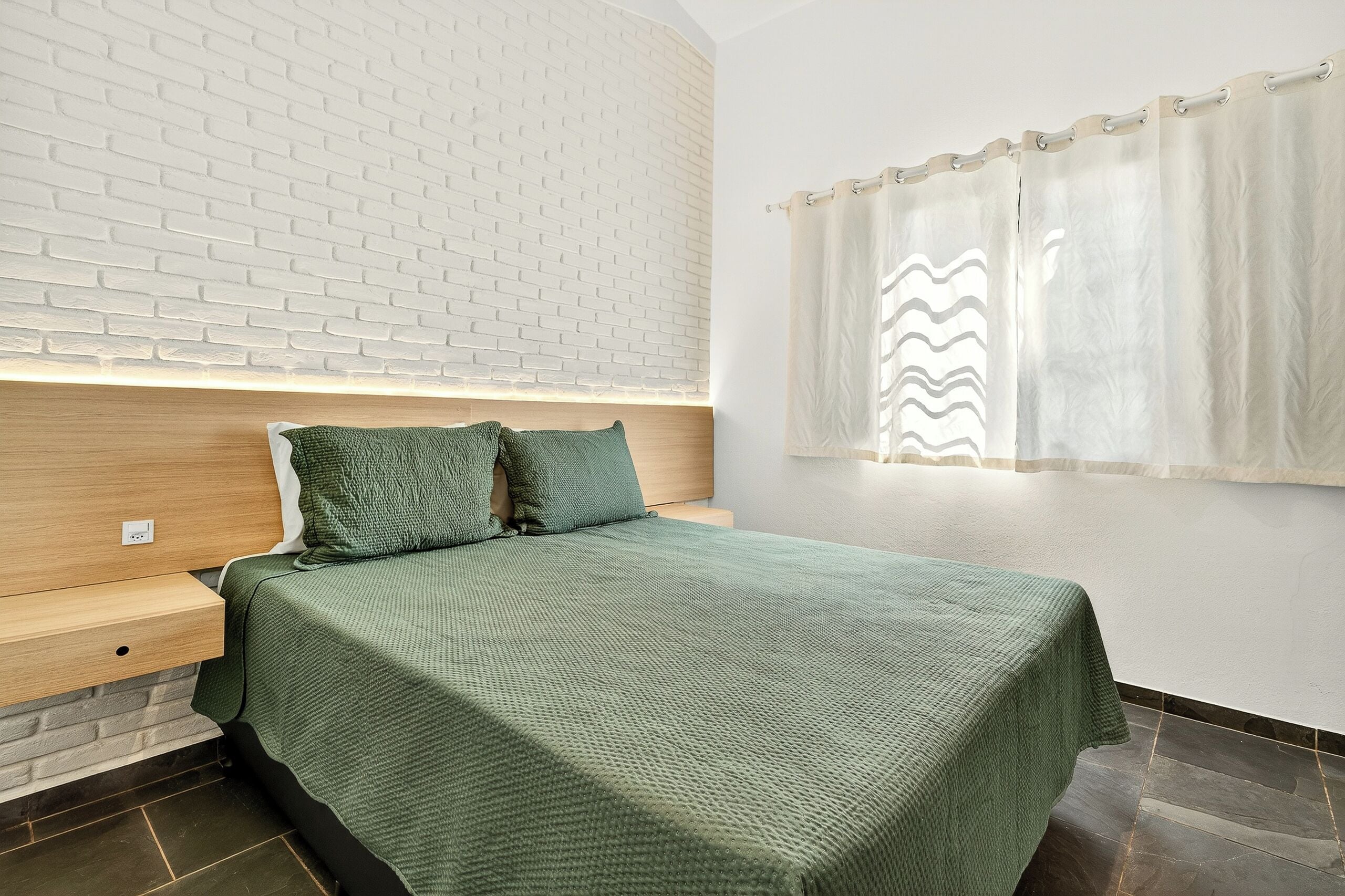 A room with a bed, a nightstand, a brick-patterned wall, a window with curtains, and a light fixture.