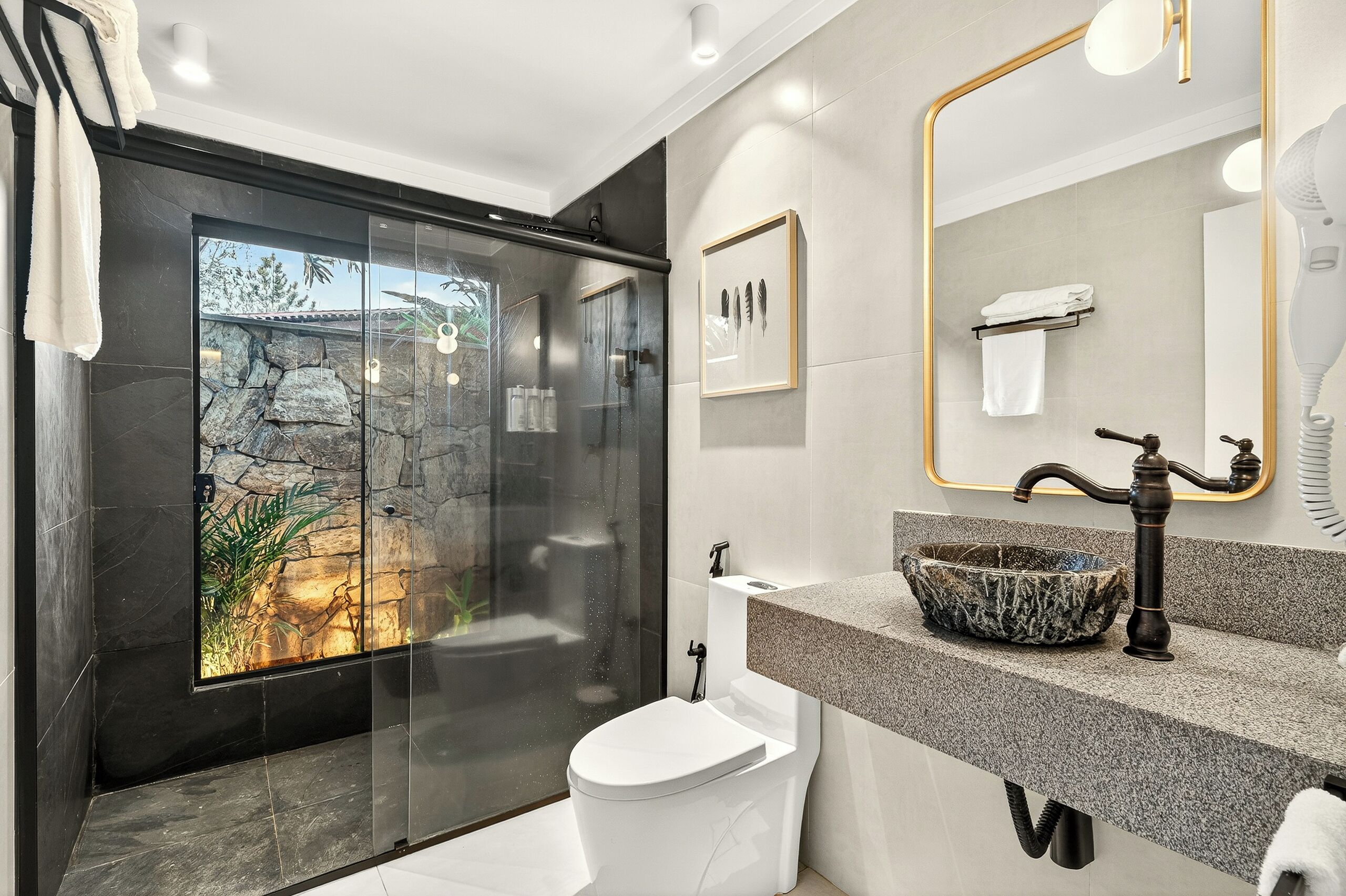 A modern bathroom with a glass-enclosed shower, a stone wall, a white toilet, a granite sink, and a large mirror with a gold frame.