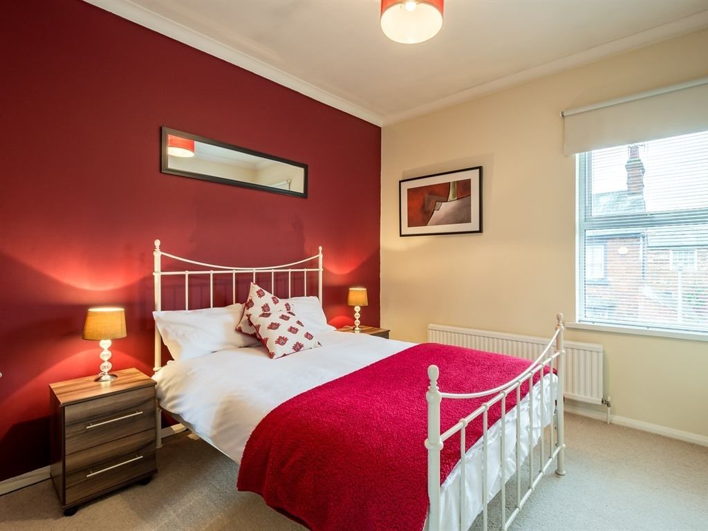 Une chambre avec un lit en métal blanc, un mur rouge et une fenêtre avec des stores.