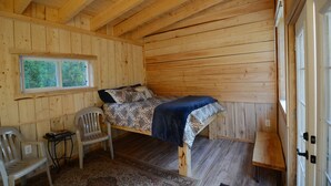 Quarto de cabana de madeira com uma cama, duas cadeiras, uma mesinha e uma janela.