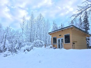 A wooden cabin with a snow-covered roof, surrounded by snow-laden trees.