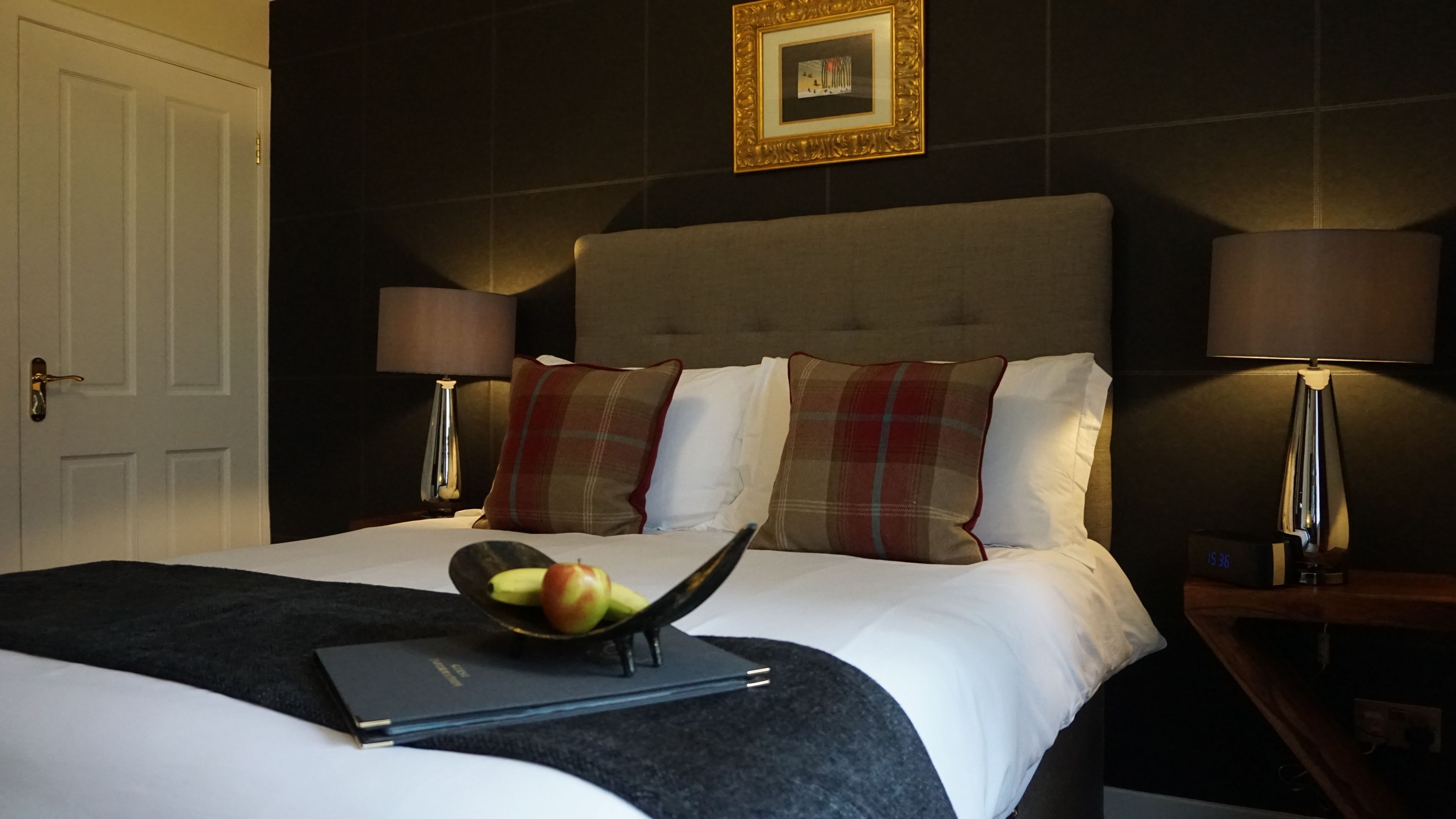 A neatly made bed with a laptop and fruit bowl on it, two bedside lamps, and a framed picture on the wall.